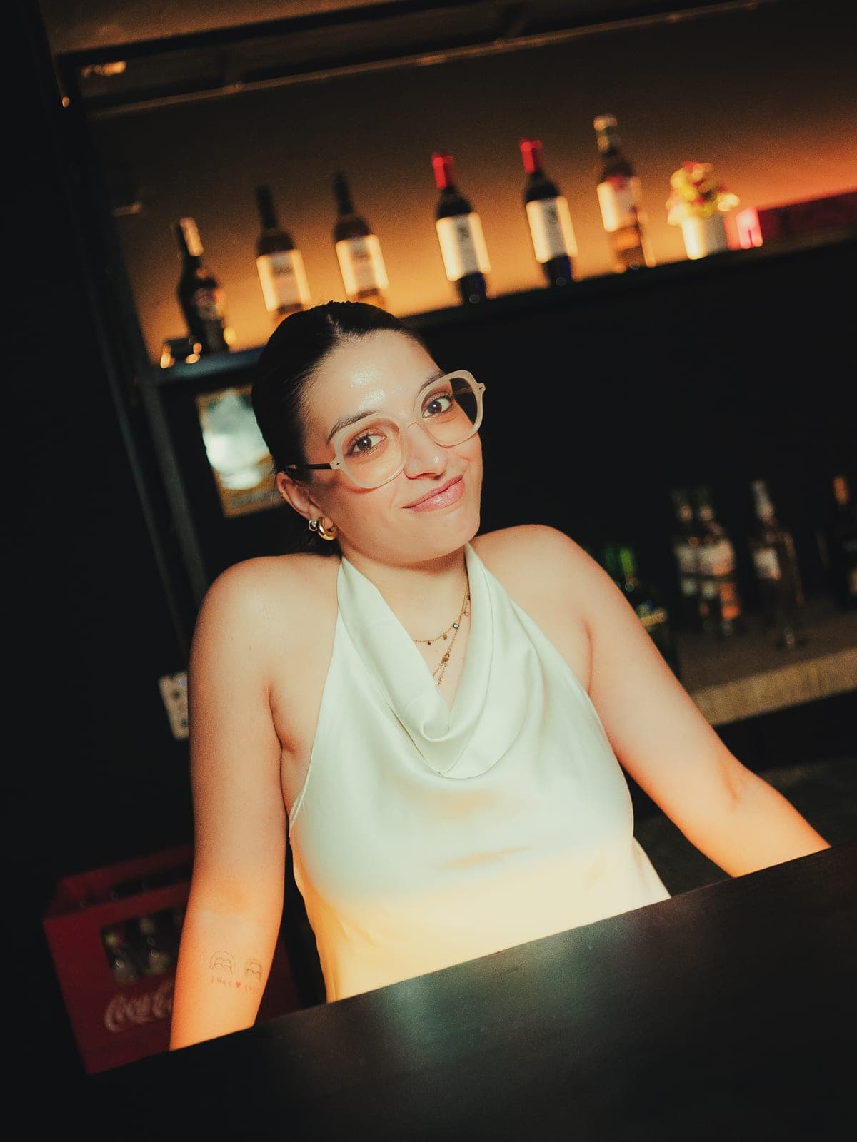 Una chica apoyada en la barra, con las botellas iluminadas detrás